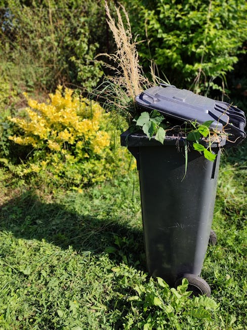 A black wheeled outdoor rubbish bin is positioned on a grassy area next to lush green bushes and trees. The lid of the bin is closed and topped with a variety of garden debris, including dried grasses