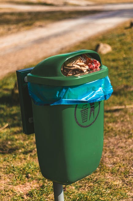 A black wheeled outdoor rubbish bin is positioned on a grassy area next to lush green bushes and trees. The lid of the bin is closed and topped with a variety of garden debris, including dried grasses